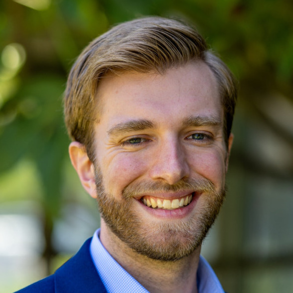 Andrew Keller's headshot. A person with short hair and a beard, wearing a collared shirt and jacket, is outside, smiling at the camera.