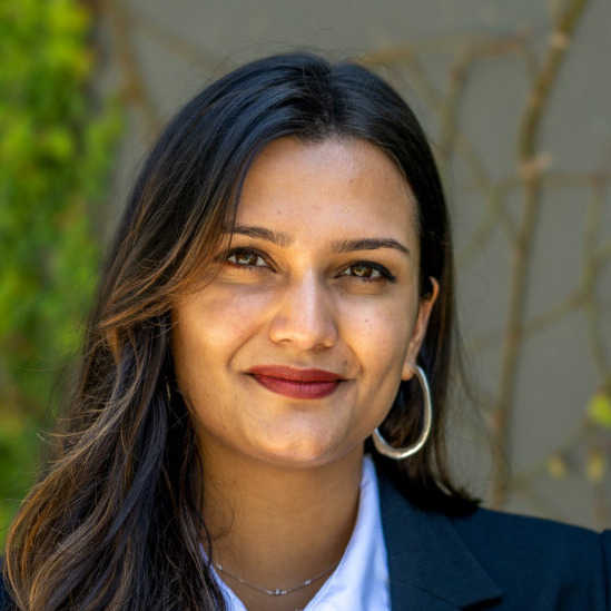 Shreya Singh Hernández's headshot. A person with long hair and side bangs, wearing a collared shirt, jacket, and hoop earrings, is outside, smiling at the camera.