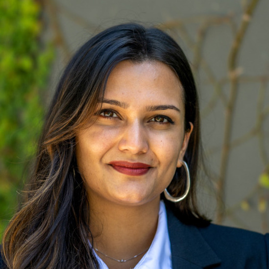 Shreya Singh Hernández's headshot. A person with long hair and side bangs, wearing a collared shirt, jacket, and hoop earrings, is outside, smiling at the camera.
