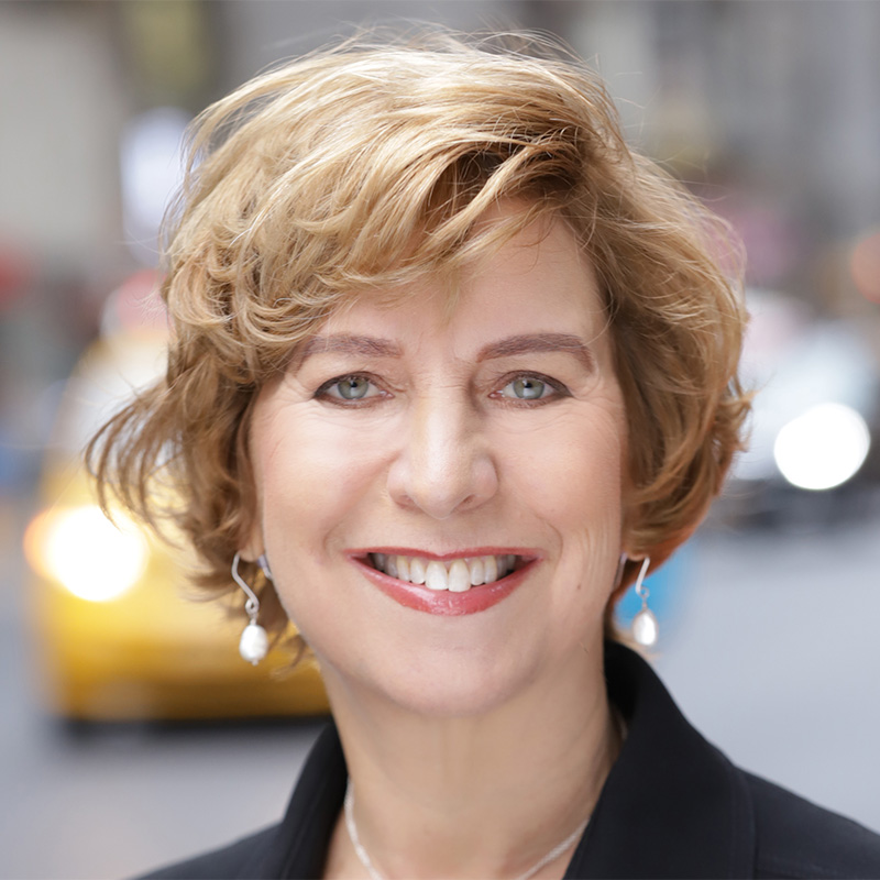 Vivian Schiller's headshot. A person with short hair, waring earrings and a jacket, is on a city street, smiling at the camera.