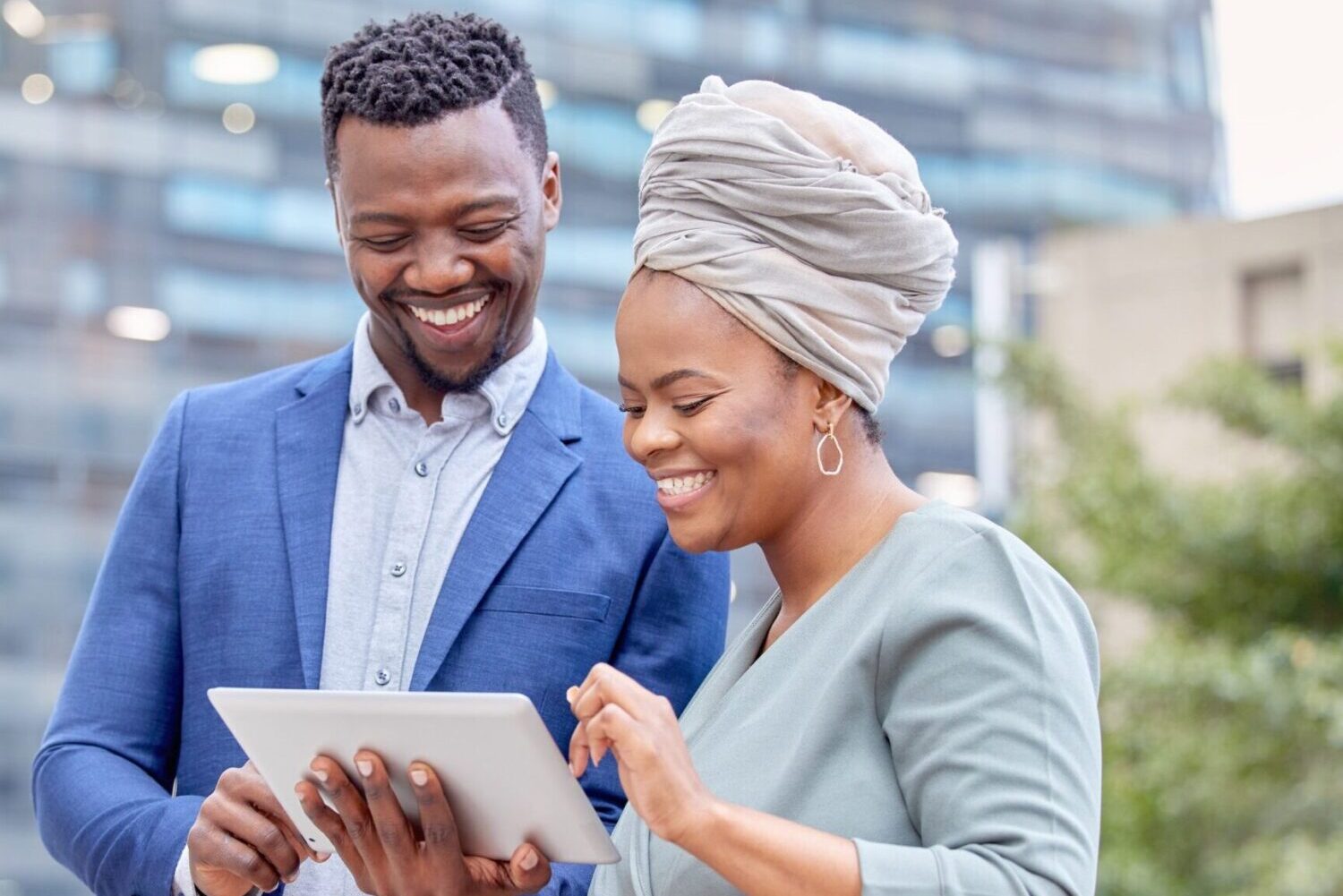A photograph of two people looking downward and smiling. The one on the left has short hair and a goatee and wears a jacket. The one on the right wears a South African head covering called a doek and hoop earrings.