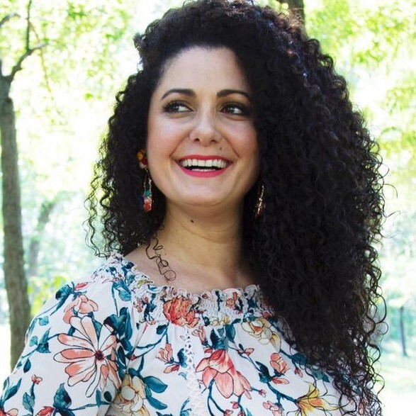Hazami Barmada's headshot. A person with long, dark, curly hair wearing a floral blouse and dangly earrings stands outside and smiles.