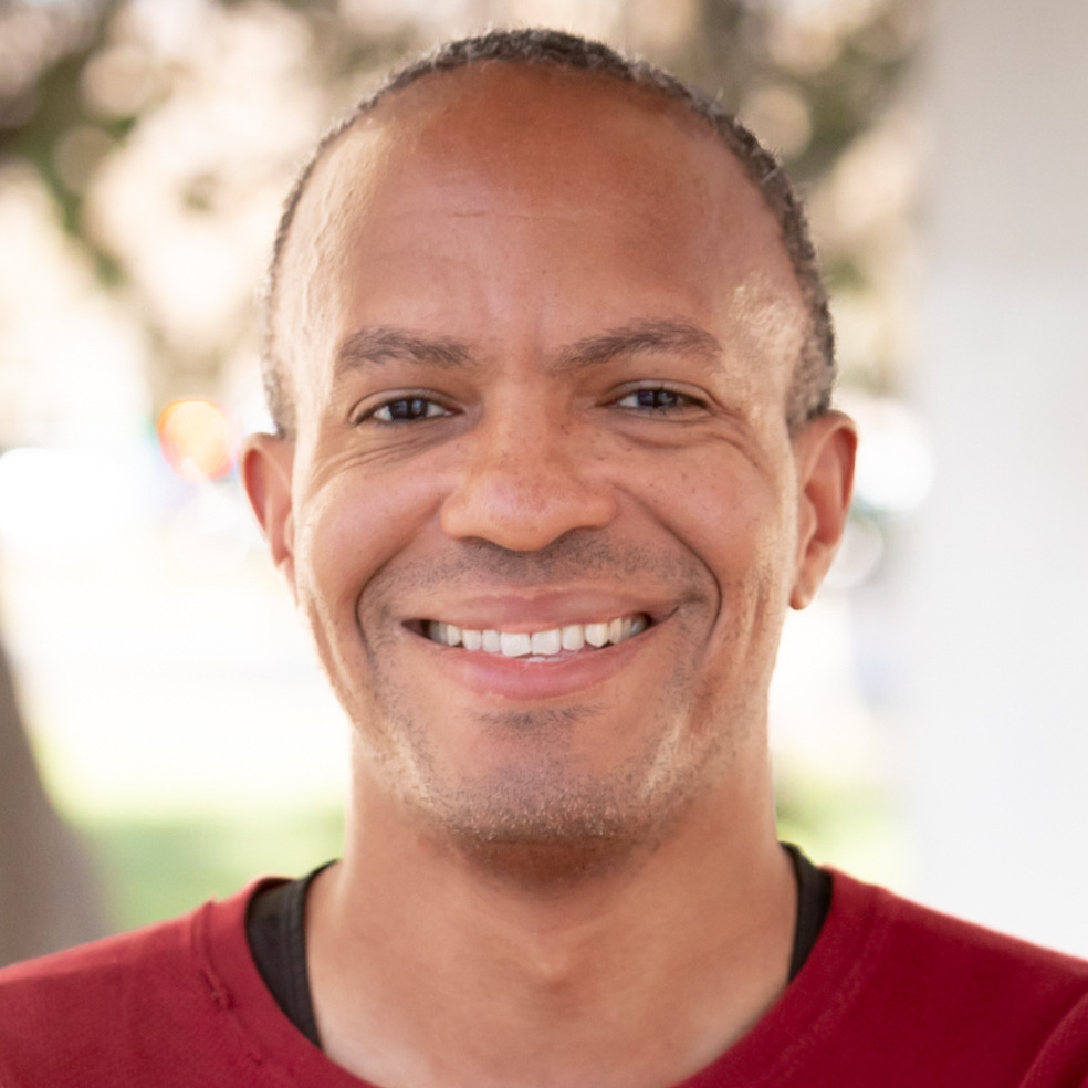 Andrew Lewis's headshot. A person with a buzzed haircut is outside, smiling at the camera.