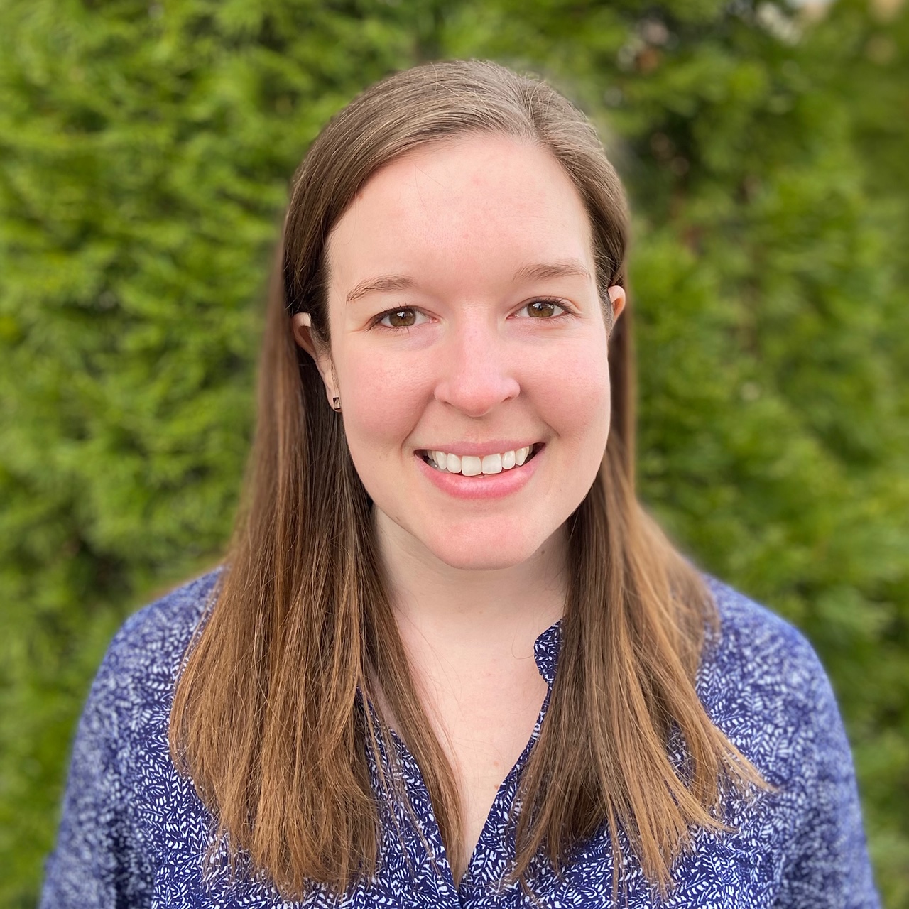 Beth Semel's headshot. A person with long hair, wearing a collared shirt, is outside, smiling at the camera.