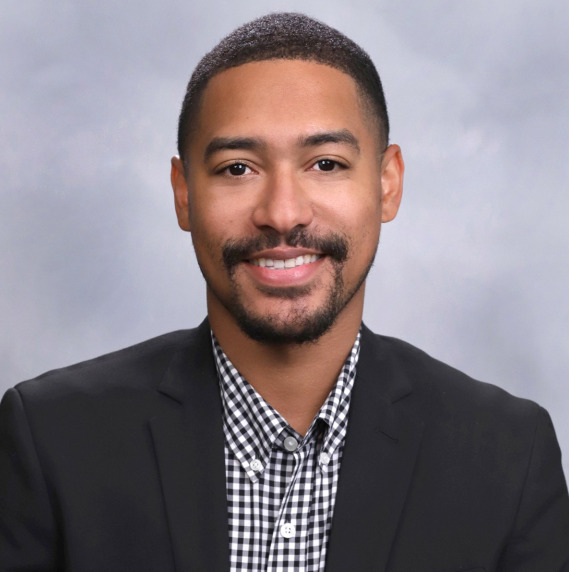 Broderick Johnson's headshot. A person with a buzzed haircut and goatee, wearing a collared shirt and jacket, smiles at the camera.