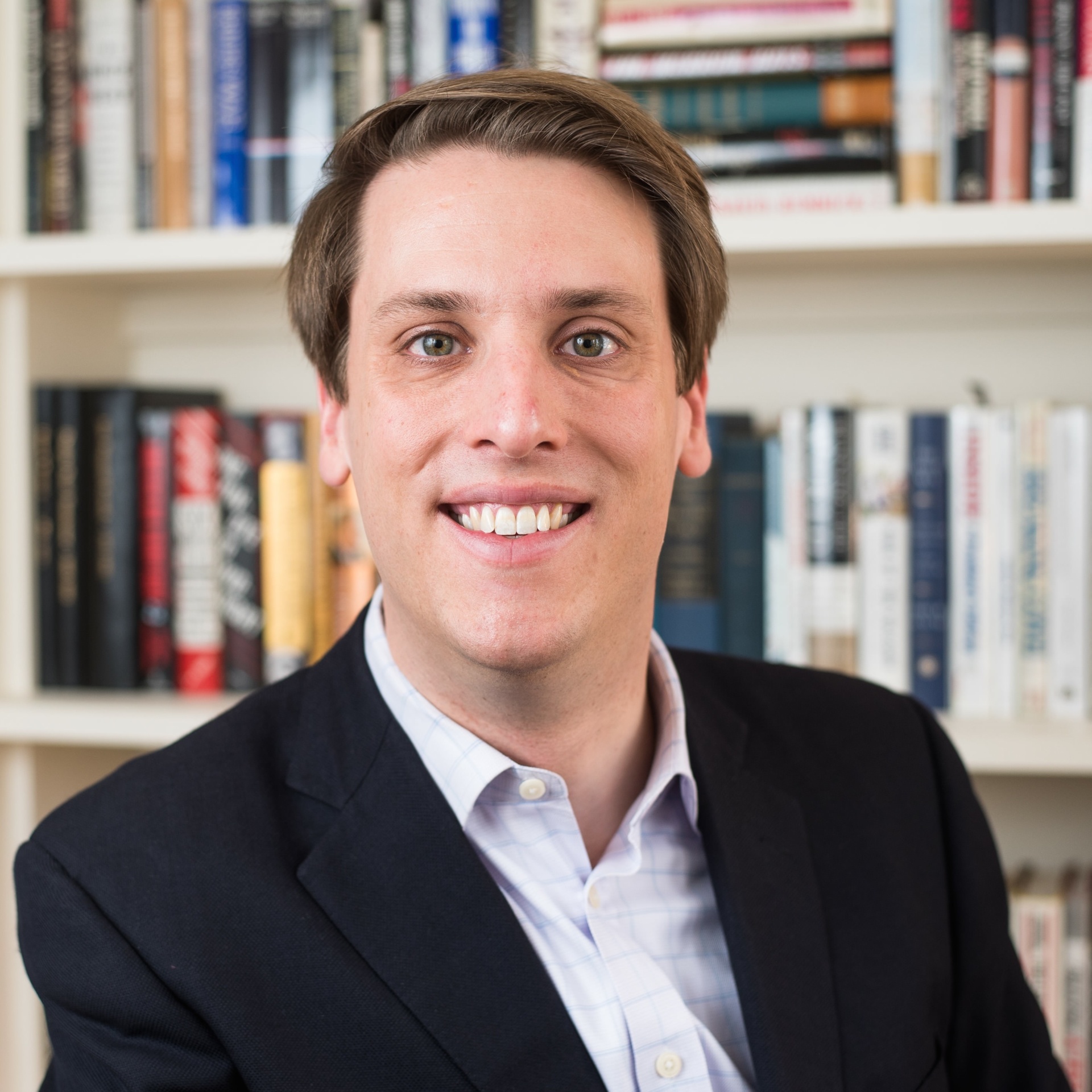 Garrett Graff's headshot. A person with short hair, wearing a collared shirt and jacket, sits in front of a bookshelf smiling at the camera.