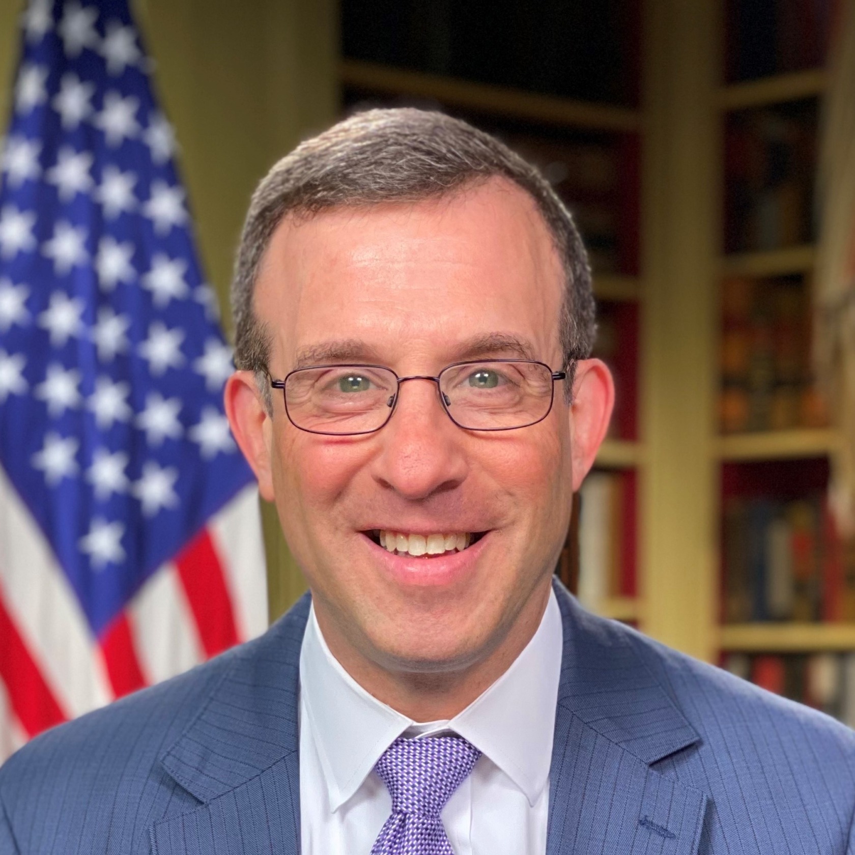 Jeff Greene's headshot. A person with short hair, wearing glasses, a jacket, and a tie, is in front of a bookshelf and American flag, smiling at the camera.