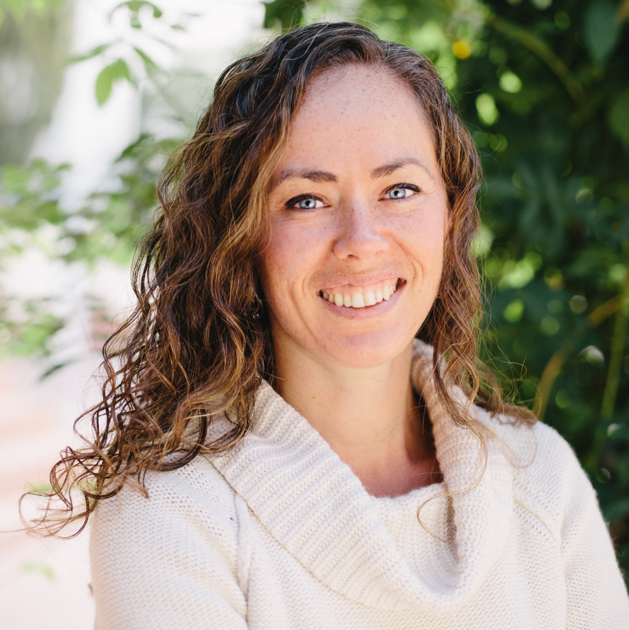 Leana Mayzlina's headshot. A person with medium-length curly hair, wearing a sweater, is outside, smiling at the camera.