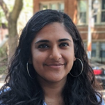 Maitreyi Sistla's headshot. A person with long, curly hair, wearing hoop earrings, is on a city street, smiling at the camera.