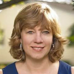 Patricia Kirsch Kelly's headshot. A person with shoulder-length hair and bangs, wearing a short-sleeved blouse and hoop hearings is outside, smiling at the camera.