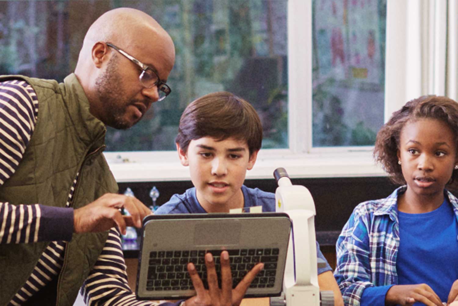 A photograph of a teacher with a tablet speaking to two young students.