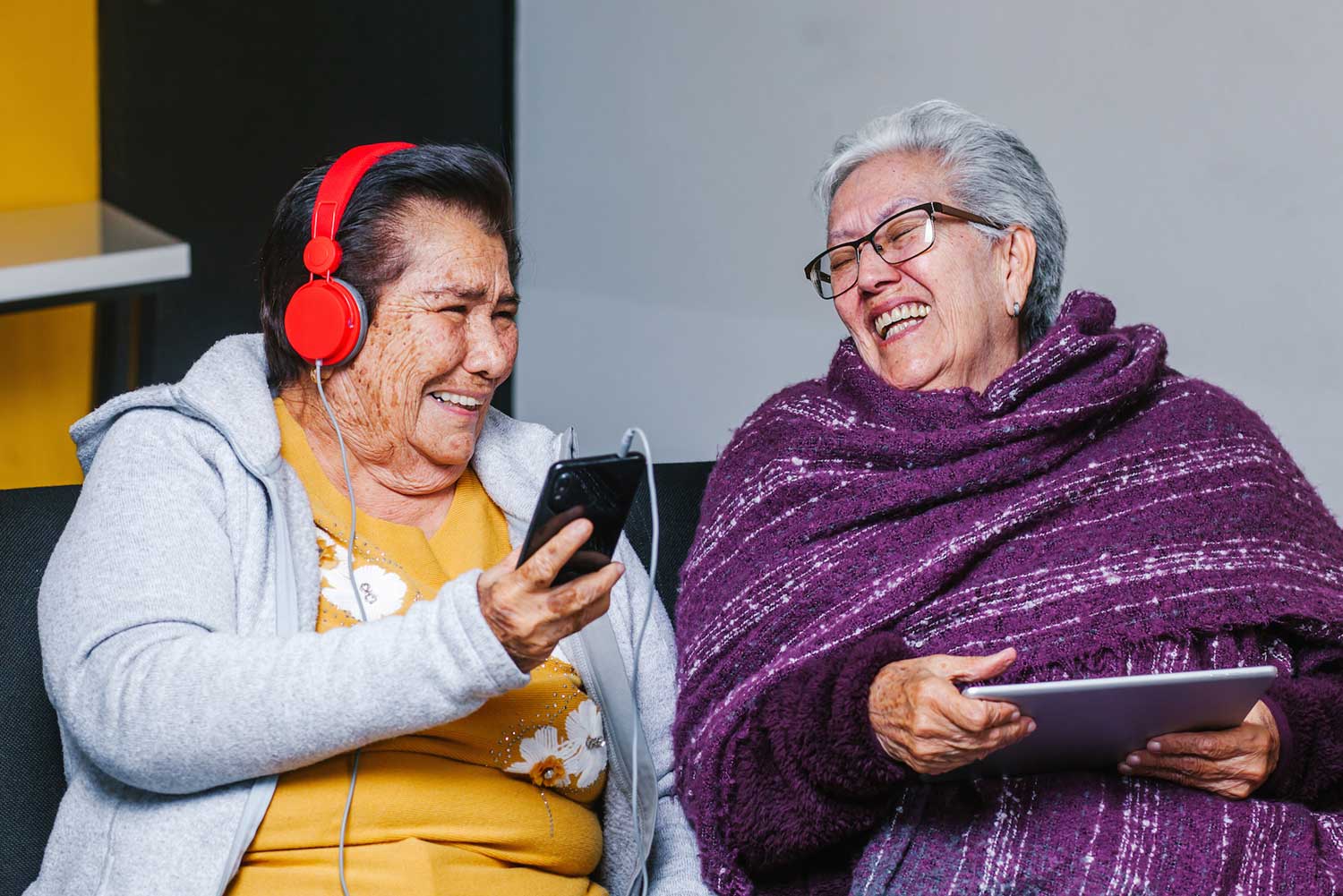 Two older adults laugh as one listens to a smartphone with headphones on and the other looks at a computer tablet.