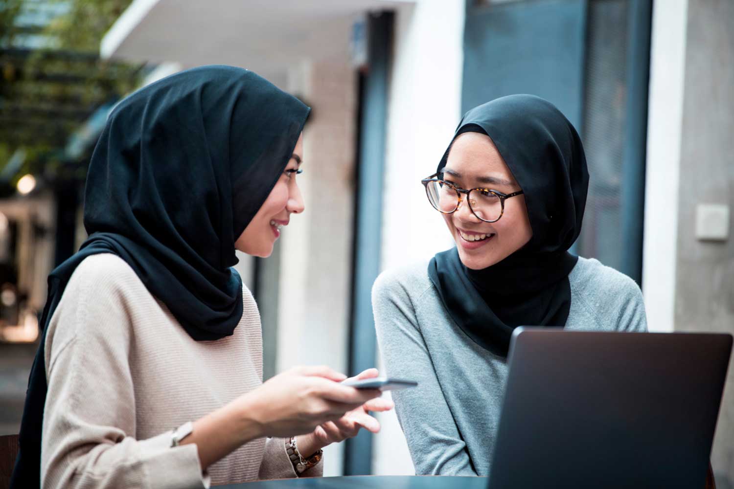 Two young people wearing head scarves sit interacting with one another, one on a smartphone and the other on a laptop computer.