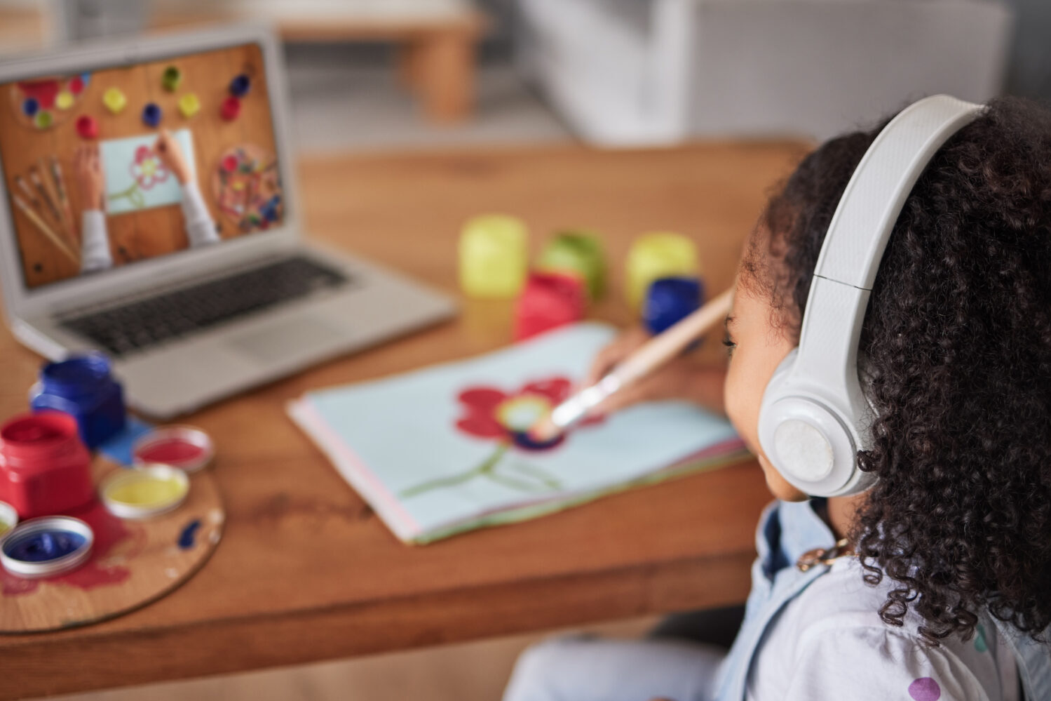 A child watches an art teacher paint a flower on their laptop screen and draws the same image.