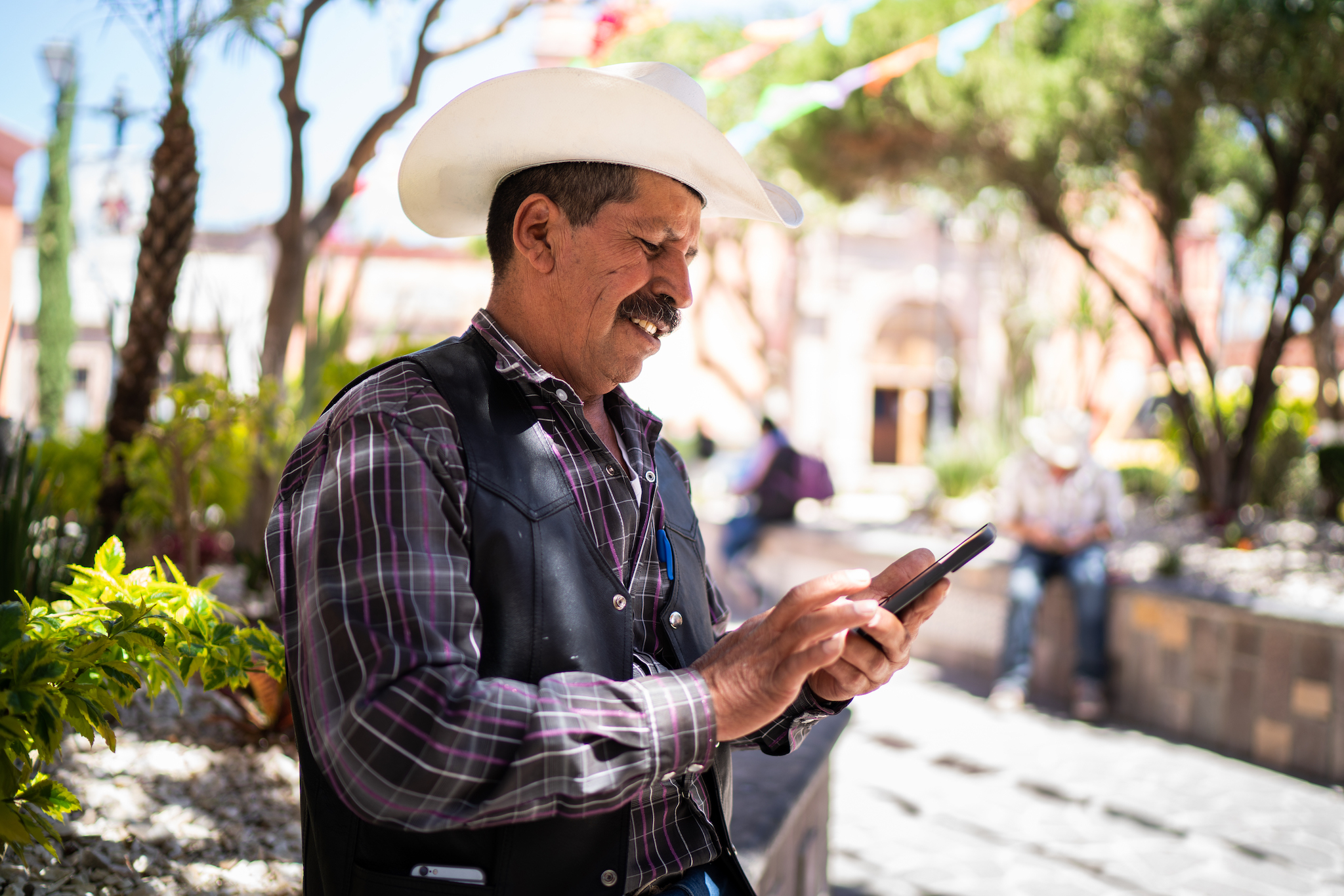 A photo of a person with short hair and a mustache wearing a cowboy hat stands outside using a smart phone.