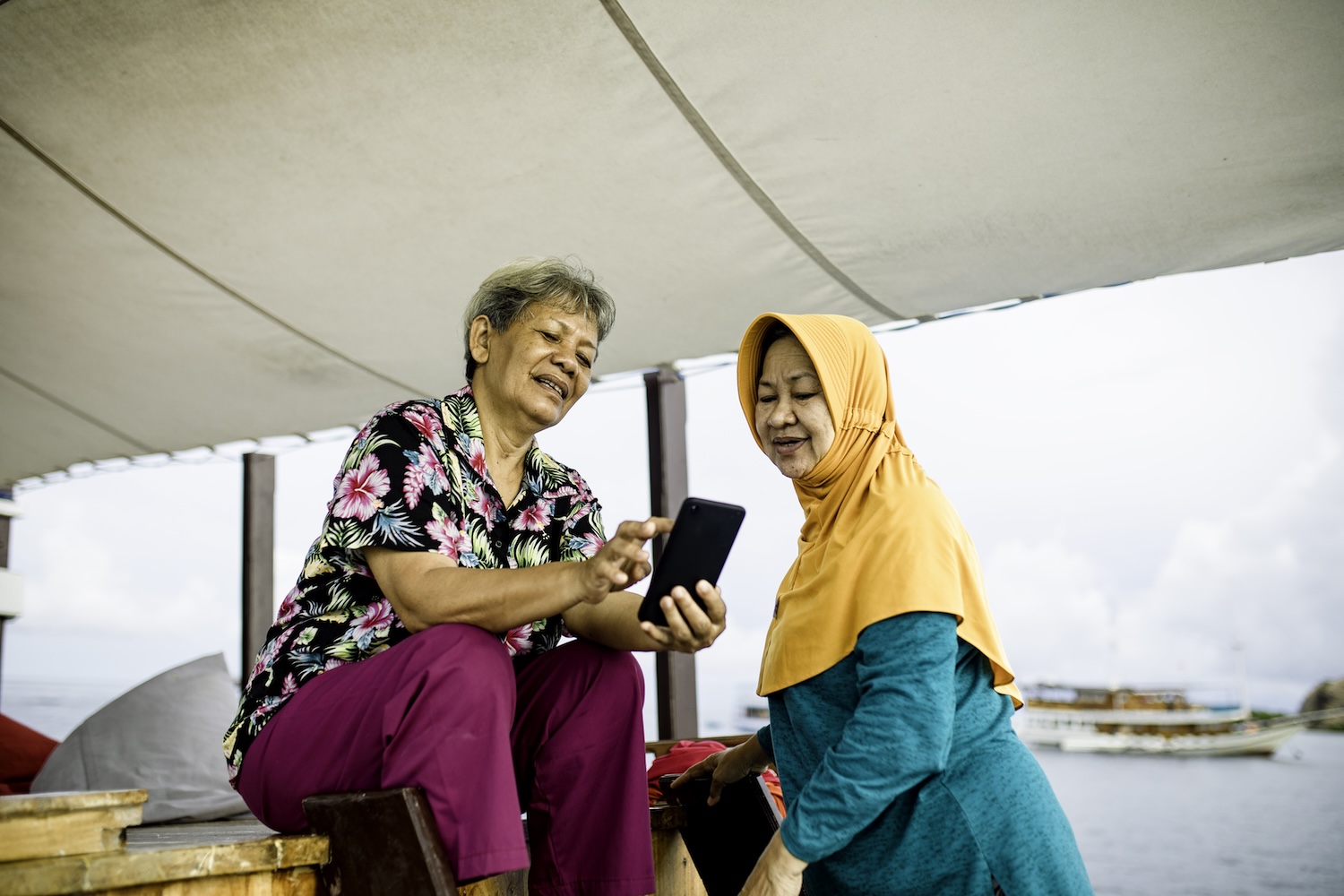 Two Malaysians look at a smart phone together. One is wearing a head covering.