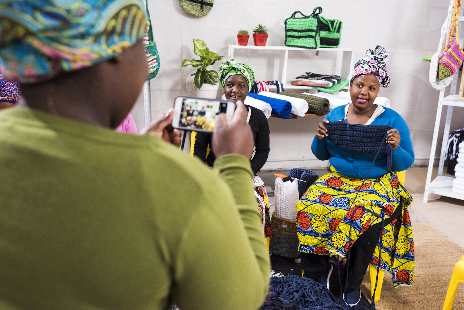 A photo of three South Africans in head coverings. One stands and takes a photo of the other two, who are seated and displaying their crocheting projects.