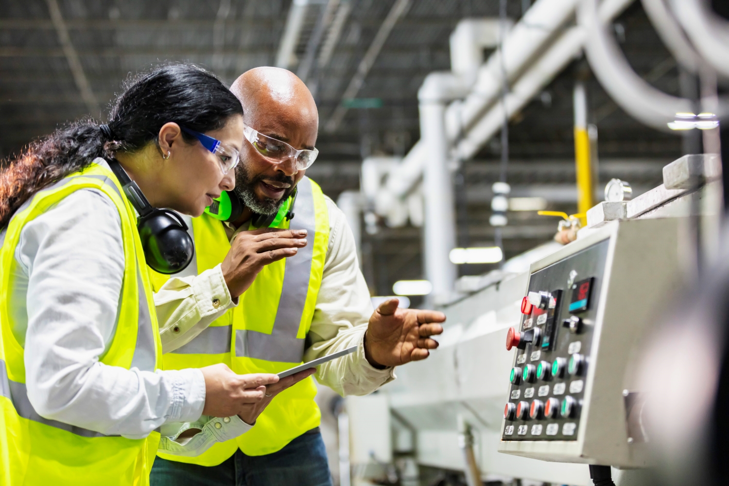 Two manufacturing workers examine equipment. One worker is holding a tablet.